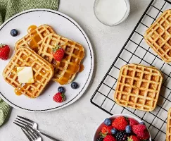 Trois gaufres de babeurre dans une assiette servies avec une noix de beurre et des fraises et des bleuets, présentées sur un comptoir de cuisine avec d'autres gaufres sur une grille de refroidissement, un verre de lait et un bol de baies mélangées.
