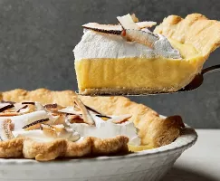 Un morceau de tarte à la crème de coco présentée de côté sur une pelle à tarte, fraîchement coupée d'une tarte entière dans un plat à tarte blanc décoratif, garnie de crème fouettée et de copeaux de noix de coco.