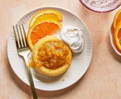 Vue en plongée d'un gâteau éponge unique nappé de marmelade d'agrumes brillante et de zeste, servi sur une assiette blanche cannelée avec des tranches d'orange fraîches et une touche de crème fouettée, accompagné d'une fourchette, sur un fond aux tons pêche doux.