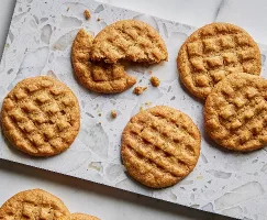 Sept biscuits au beurre d'arachide dorés avec des motifs distinctifs faits à la fourchette présentés sur du papier parchemin sur une surface en marbre, l'un partiellement mangé pour révéler une texture douce et moelleuse.