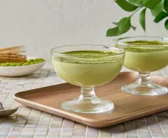 Deux tasses en verre de mousse au chocolat blanc et matcha garnies de poudre de matcha verte sur un plateau en bois, avec un fouet à matcha traditionnel et un bol de poudre de matcha en arrière-plan, posés sur une table carrelée à la lumière naturelle.