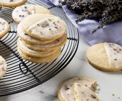 Une fournée de sablés citron-lavande glacés saupoudrés de lavande séchée disposés sur une grille de refroidissement circulaire sur un comptoir en marbre, avec des tiges de lavande séchée et un linge couleur lavande