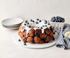 Monkey bread estival aux bleuets sur une assiette blanche avec du glaçage blanc coulant et des bleuets frais parsemés sur le dessus, disposé à côté d’une serviette rayée bleue et blanche, d’un bol bleu contenant du glaçage et d’un panier de bleuets, le tout sur une surface de couleur pâle.