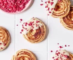 Vue aérienne de biscuits à la pâte feuilletée aux framboises avec glaçage au chocolat blanc et morceaux de framboises lyophilisées sur un comptoir carrelé blanc, avec un bol de framboises écrasées.
