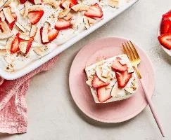 Une part de gâteau tres leches aux fraises et à la noix de coco sur une assiette rose avec une fourchette dorée, déposée à côté d’un moule blanc contenant le même gâteau et d’un petit bol de fraises tranchées, le tout sur un comptoir gris avec une serviette en tissu rose.
