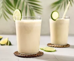 Deux verres de limade brésilienne crémeuse sont garnis de quartiers de lime fraîche et reposent sur des sous-plats tissés à la main. Les breuvages sont posés sur une surface de carreaux pâles, avec des palmiers en arrière-plan. D'autres quartiers de lime sont éparpillés sur la table.
