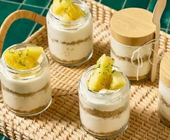 Un plateau en osier, adossé à un arrière-plan de carreaux verts, contient plusieurs verrines de cheesecake à l'ananas en portions individuelles. Chaque verrine présente des couches de gâteau au fromage crémeux et de croûte friable au graham cracker, garnies de morceaux d'ananas frais et d'une pincée de zeste de lime. Deux des verrines ont des couvercles en bois, et une d'entre elles est accompagnée d'une cuillère en bambou attachée