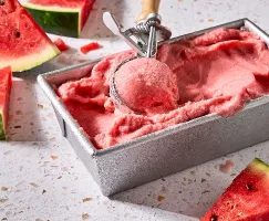 Un contenant en métal rempli de sorbet crémeux à la pastèque, en train d'être portionné avec une cuillère à crème glacée vintage. Des tranches de pastèque fraîche sont dispersées autour du contenant sur un comptoir en calcaire de couleur claire.