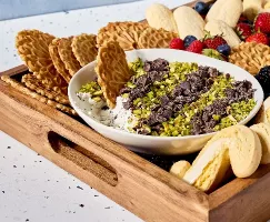 Un plateau en bois rempli d'un assortiment de biscuits italiens et de fruits frais - des fraises, des bleuets et des mûres - entoure un bol de trempette à cannoli. La trempette est garnie de pistaches hachées et de morceaux de chocolat, et on y voit un biscuit Pizzelle en train de tremper.