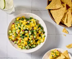 Une image en plongée d'un bol de salsa à la mangue et au concombre sur une surface en carreaux blancs. La salsa comprend également de l' oignon rouge et de la coriandre. Un bol de chips tortilla est placé à côté, avec quelques chips éparpillés autour. On voit également un verre d'eau infusée à la lime.