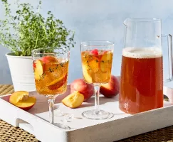Un plateau blanc posé sur une table en osier supporte deux verres à pied, un pichet de thé sucré à la pêche et au thym, ainsi que des pêches fraîches entières et coupées. Les boissons sont garnies de tranches de pêches fraîches et de brins de thym. On aperçoit également un plant de thym en pot en arrière-plan.