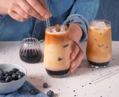  Une personne en chemise de jeans brasse un café glacé aux bleuets avec une paille en verre. La boisson a des couches distinctes de sirop de bleuets, de lait et de café. Une deuxième boisson identique est posée à proximité sur un comptoir moucheté. Un pichet en verre de sirop de bleuets, un bol de bleuets frais et des bleuets éparpillés complètent la scène.