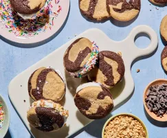 Un plateau de sandwichs à la crème glacée marbrée au chocolat et à la vanille est posé sur une planche de service blanche. Certains sont roulés dans des vermicelles de couleur ou des noix hachées. D'autres biscuits et des bols de garnitures entourent la scène sur une surface bleu pâle.