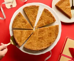 Part de gâteau néerlandais au beurre et à la cassonade posée sur un support à gâteau blanc sur fond rouge. Une des parts est prélevée à l’aide d’une pelle à gâteau dorée. Une autre part est posée sur une petite assiette blanche accompagnée d’une fourchette dorée, et un paquet-cadeau orné d’un ruban se trouve juste à côté.