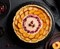 Tarte à la crème aux prunes décorée de tranches de prunes et de pétales de roses séchées sur un plateau noir déposé sur une tabl