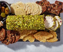 Une bûche au fromage à la crème enrobée de pistaches concassées sur une planche avec croustilles, craquelins et olives