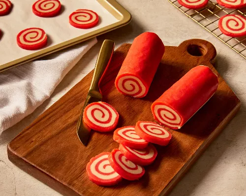 Deux rouleaux de pâte à biscuits en spirale aux couleurs rouge et blanche, posés sur une planche à découper en bois avec plusieurs tranches coupées, et d'autres tranches sur une plaque de cuisson, ainsi que des biscuits cuits refroidissant sur une grille en métal